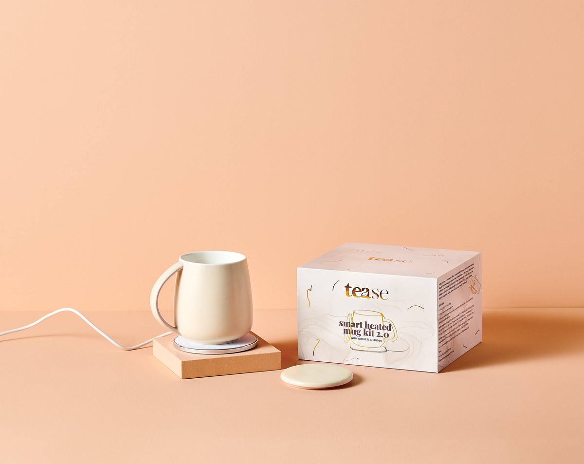 A white ceramic smart heated mug kit set on a wooden base next to its white packaging box, with a matching white coaster in front, all set against a solid peach background.