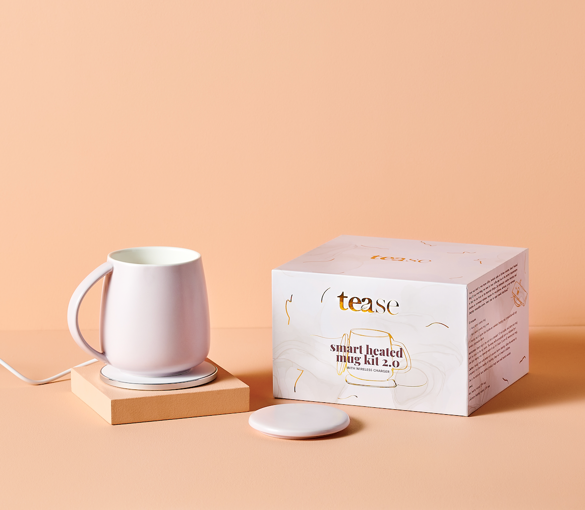A smart heated mug kit by tease, featuring a white ceramic mug on a wooden heating base, a white wireless charging pad, and its white packaging box with gold accents, set against a solid peach background.